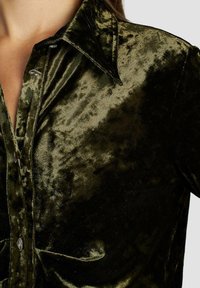Camisa de terciopelo verde oscuro con una textura suave, cierre de botones y cuello puntiagudo. Presenta botones metálicos brillantes y variaciones sutiles en el brillo.