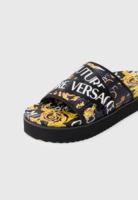 Sandália preta de calçar com parte superior estampada com padrão de arabescos dourados e texto branco. Sola espessa e texturizada com design minimalista.