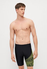 Μαύρα μαγιό σορτς με εφαρμοστό σχέδιο, που διαθέτουν ένα ζωντανό κίτρινο γεωμετρικό μοτίβο στη μία πλευρά. Κατασκευασμένα από λείο, ελαστικό υλικό.