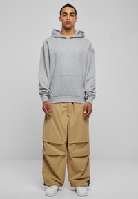 Felpa grigia oversize con tasca frontale, abbinata a pantaloni beigi larghi con design a strati. Le sneakers bianche completano l'outfit.