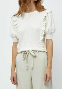 Vit kortärmad topp med volanger på axlarna, rund halsringning och avslappnad passform. Kombinerad med högmidjade ljusa byxor.