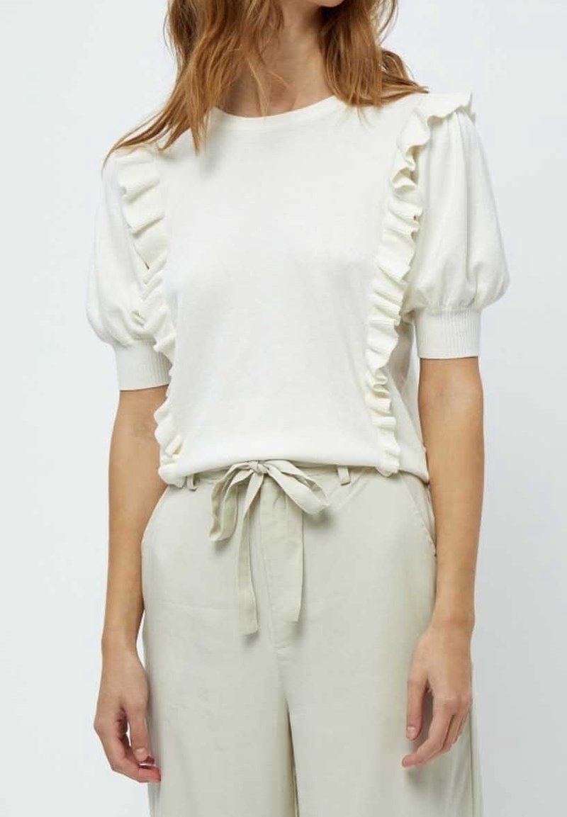 Vit kortärmad topp med volanger på axlarna, rund halsringning och avslappnad passform. Kombinerad med högmidjade ljusa byxor.