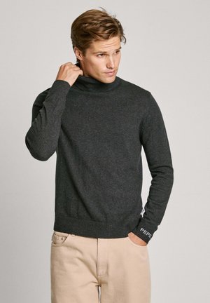 Suéter de cuello alto en gris oscuro, elaborado en tejido de punto, con puños y dobladillo acanalados, que presenta un detalle del logo en color blanco en el puño.