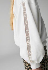 Vit sweatshirt med lösa ärmar, prydd med en beige rand längs ärmen med präglad text "01-11-94 ALLSAINTS." Texturerat tyg; avslappnad passform.