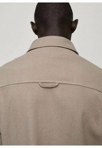 Beige jas met een klassieke kraag en gestikte schouderpas aan de achterkant. De stof heeft een textuurafwerking en een lus aan de achterkant om op te hangen.