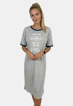 Mujer con coleta rubia, vestida con un vestido gris hasta la rodilla con ribete oscuro y texto que dice "Bring me coffee and tell me I'm pretty", de pie con las manos entrelazadas.
