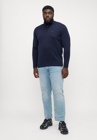 Granatowy sweter z suwakiem przy kołnierzu, w zestawie ze jasnoniebieskimi dżinsami. Model nosi sneakersy w kolorze czarnym i fioletowym. Prosty, dopasowany krój.
