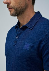 Tummansininen polo-paita, jossa on teksturoitu kangas, kahden napin läppä ja neliömäinen SALSA JEANS -logo-merkki vasemmassa rinnassa.