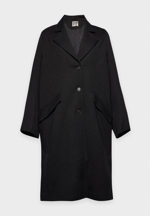 Une femme porte un long manteau noir sur un col roulé couleur crème, un pantalon olive et des chaussures noires, avec un sac foncé en bandoulière.