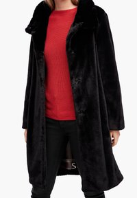 Černý plášť z umělé kožešiny s širokým límcem, uzávěrem na knoflíky a dlouhými rukávy, nošený přes červený texturovaný svetr a černé kalhoty.