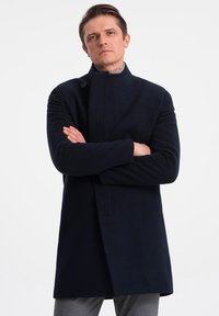 Laine navy long manteau avec un col haut, fermeture à un bouton et poches latérales. Texture lisse, coupe ajustée et design minimaliste.