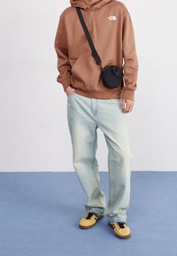 Sudadera marrón con un bolsillo frontal, jeans de mezclilla azul claro y zapatillas negras y amarillas. Una pequeña bolsa cruzada negra cuelga del hombro.