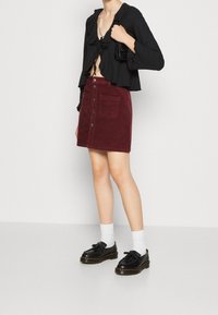 Persona con un top corto negro de mangas largas, falda burdeos con botones delanteros y bolsillos, calcetines blancos, mocasines negros y sosteniendo un bolso negro.