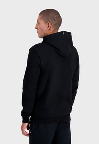 Sudadera negra con capucha y cordón, con puños acanalados, hecha de un tejido suave, que cuenta con una pequeña etiqueta de logo en la parte posterior. Diseño simple y estilizado.