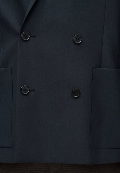 Μπλε σκούρο διπλόμπουκο σακάκι από λείο ύφασμα, με δύο μαύρα κουμπιά και δύο μπροστινές τσέπες. Μινιμαλιστικός σχεδιασμός με αιχμηλές πέτες.