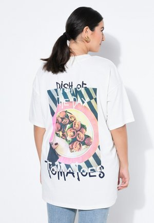 Nainen, jolla on tummat poninhännät, pukeutuneena valkoiseen ylisuurikokoiseen T-paitaan, jossa on kuva lautasesta, jossa on tomaattisella paahdolla koristeltu leipä ja teksti "Päivän ruoka Tomaatit" takana.