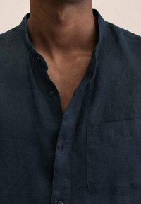Camicia in lino blu navy con colletto alla mandarino, una tasca sul petto e chiusure con bottoni. Il tessuto texturizzato aggiunge profondità al design.