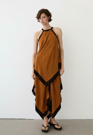 Kobieta w bezrękawnej sukience w kolorze rdzy z czarnym wykończeniem, asymetrycznym dołem i czarnych sandałach na obcasie, stojąca przy białej ścianie.