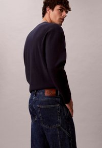 Maglione blu marino a maniche lunghe con scollo a costine abbinato a jeans in denim blu scuro con toppa con logo in pelle e multiple tasche.