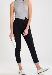 Grå ärmlös topp, marinblå skräddarsydda byxor med ränder och knytmidja, samt vita slip-on skor; håller en vit clutch.