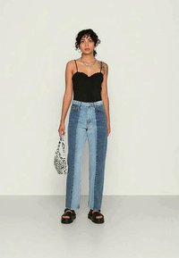 Mulher com cabelo encaracolado, usando uma camisola preta sem mangas, jeans bicolores azuis, sandálias de plataforma pretas, colar prateado e segurando uma pequena bolsa estampada.
