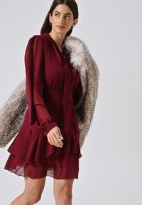 Burgunderfarbnes Chiffonkleid mit einem Rüschensaum, langen Ärmeln und einem Bindekragen; kombiniert mit einem leichten Pelzmantel. Verfügt über silberne Ringaccessoires.