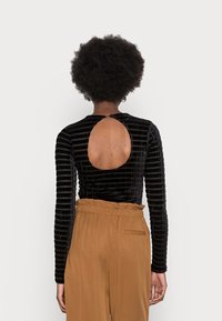 Schwarzes Samtoberteil mit vertikalen Streifen und einem runden Ausschnitt im Rücken, kombiniert mit braunen, hoch taillierten Hosen.