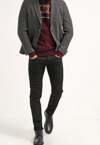 Blazer gris porté sur un pull bordeaux avec des accents à motifs, associé à un pantalon noir ajusté et des chaussures noires, présentant une texture lisse et une coupe sur mesure.