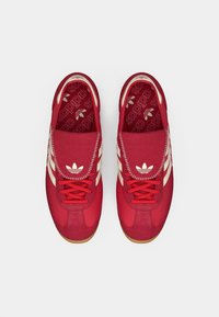 Sneakers rosse con tomaia testurizzata, caratterizzate da strisce color crema e lacci, una linguetta imbottita e suola in gomma marrone. L'interno presenta un motivo con il logo.