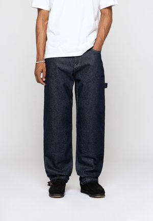 Homme portant un jean bleu foncé ample, un t-shirt blanc, des chaussures noires, debout avec une main dans la poche devant un fond uni.