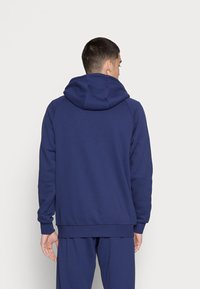 Marineblå hættebluse med en stor, justerbar hætte, ribbede manchetter og kant. Glat stof med en afslappet pasform, set fra bagsiden.