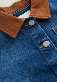 Džinsinė striukė su rūdžių spalvos kordurovo apykakle ir metalinėmis spaudėmis, rodanti išsamią tekstūrą ir siuvimą.