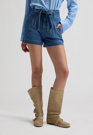 Person iført højtaljede denimshorts med et bundet bælte, lyseblå langærmet skjorte og beige knæhøje støvler på en ensfarvet baggrund.