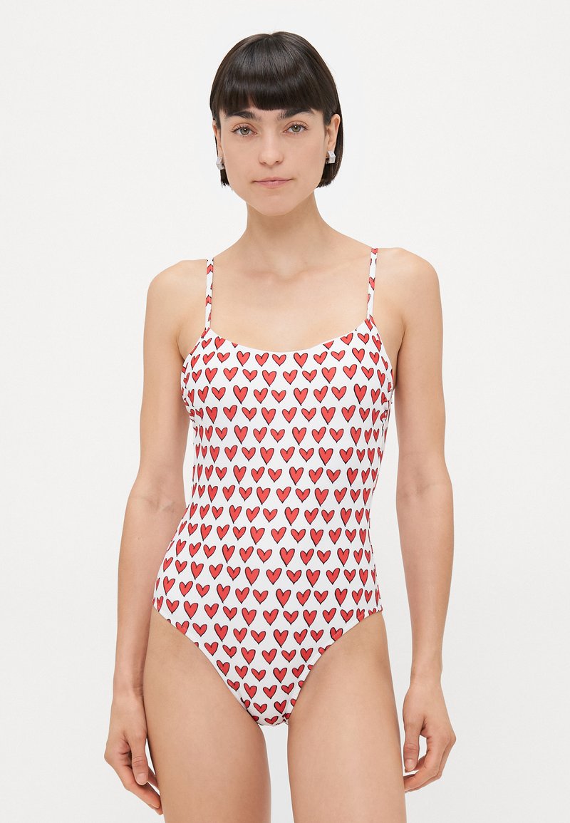 One-piece swimsuit featuring a white base with a repeating pattern of red hearts, adjustable thin straps, and a classic scoop neckline.