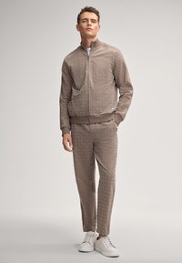 Man in een bruine, gemêleerde ritssluitingjacket en bijpassende broek met witte sneakers, staand tegen een effen lichte achtergrond.