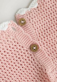 Tissu tricoté rose avec un motif à armure ouverte. Comprend une bande texturée, des accents blancs et des boutons ronds dorés avec gravure.