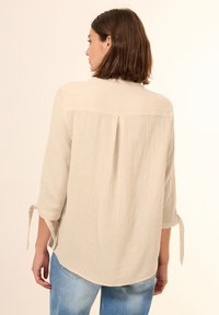 Beige, leichtes Blusenoberteil mit Dreiviertelärmel, Rückenfalte und gebundenen Manschetten, kombiniert mit hellblauen Jeans.