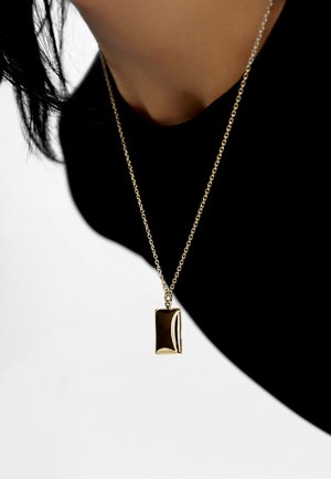 Gold-tone rectangular pendant on a delicate chain necklace, featuring a smooth, reflective surface and subtle curvature, worn against a dark top.