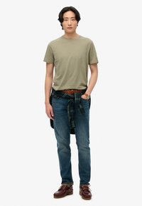 Hverdagsoutfit bestående af en lys olivengrøn t-shirt, blå jeans, en ternet skjorte knyttet om livet og brune lædersko med snørebånd.