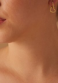 Boucles d'oreilles en forme de cœur en or suspendues à un petit fermoir, mettant en avant une finition lisse et un design minimaliste sur le teint de la peau.