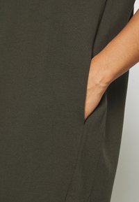 Ciemnozielona sukienka z miękkiej bawełny, z boczną kieszenią i luźnym krojem. Tkanina ma gładką teksturę i minimalistyczny design.