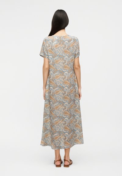 Γυναίκα με μακριά σκουρόχρωμα μαλλιά, που φοράει φόρεμα με κοντά μανίκια και μήκος μέχρι τους αστραγάλους, με μπλε και καφέ παϊένσα μοτίβο, στέκεται γυρισμένη προς τα πίσω.