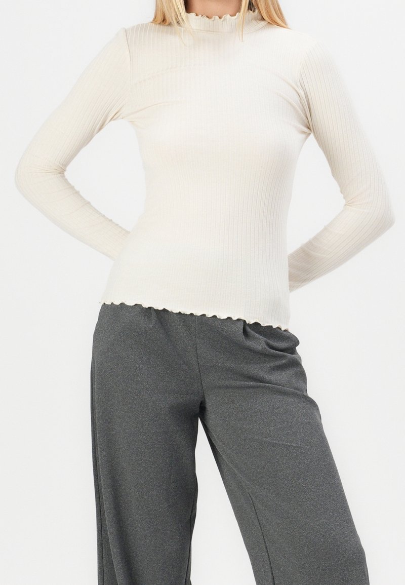 Femme portant un haut moulant côtelé crème à manches longues avec un bord en forme de laitue et un pantalon ample gris foncé, debout, les mains dans le dos.
