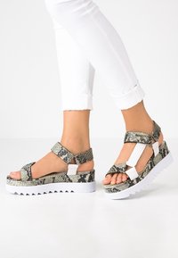 Nohy v bílých kalhotách a na platformových sandálech s hadím vzorem a bílými popruhy na bílém pozadí.