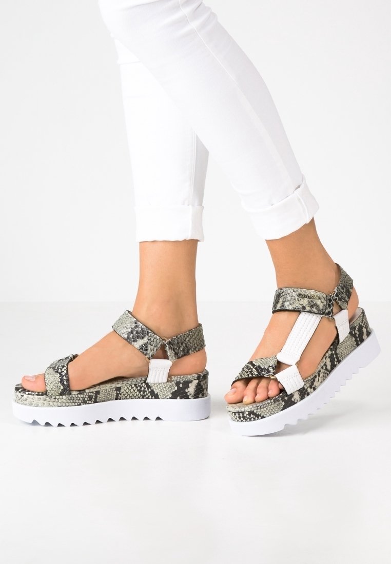 Nohy v bílých kalhotách a na platformových sandálech s hadím vzorem a bílými popruhy na bílém pozadí.