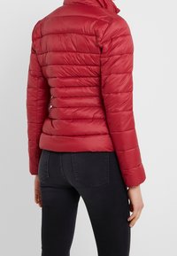 Rode pufferjack met verticale doorgestikte structuur, hoge kraag en ritssluiting. Gecombineerd met donkere jeans, waardoor een aangesloten silhouet wordt getoond.