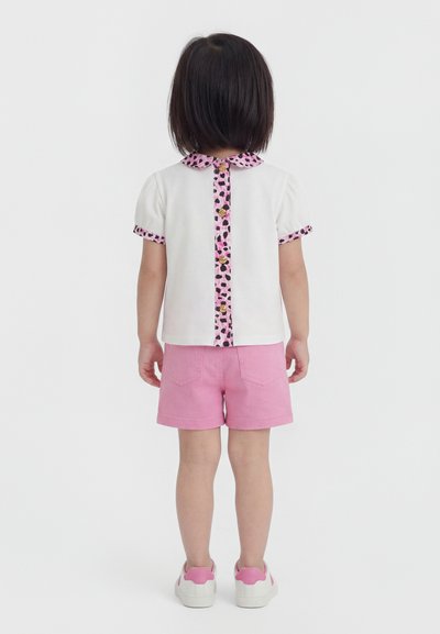 Παιδί με κοντά σκουρόχρωμα μαλλιά στέκεται με πλάτη, φοράει λευκό πουκάμισο με ροζ λεοπάρ διακόσμηση και κουμπιά, ροζ σορτς και λευκό-ροζ αθλητικά παπούτσια.