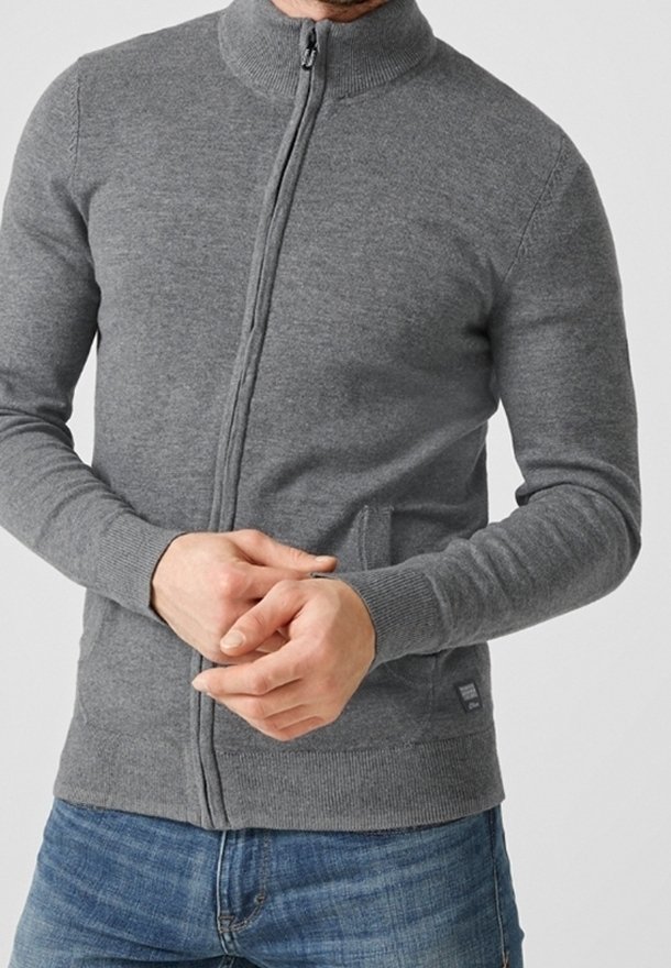 Homme portant un pull gris ajusté avec fermeture éclair, manches et ourlet côtelés, assorti à un jean bleu, mains jointes devant.