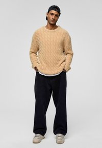 Beige kabelgebreide trui met een losse pasvorm, gecombineerd met donkere wijde broekspijpen en grijze sneakers, gedragen met een zwarte pet.