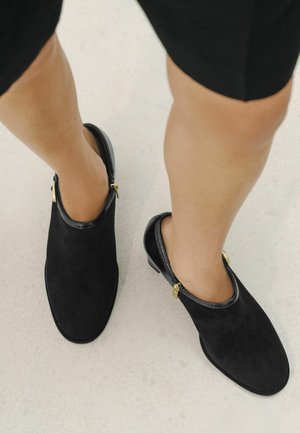 Personne portant des chaussures slip-on en daim noir avec des fermetures éclair dorées, debout sur un sol clair.